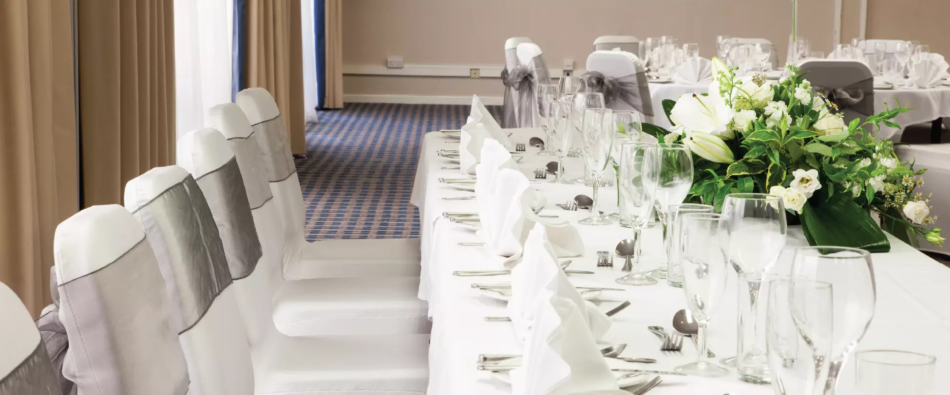 A table set up for the wedding party at a wedding breakfast. The table and chairs have white coverings and the chairs have silver ribbons. There is a floral centrepiece..
