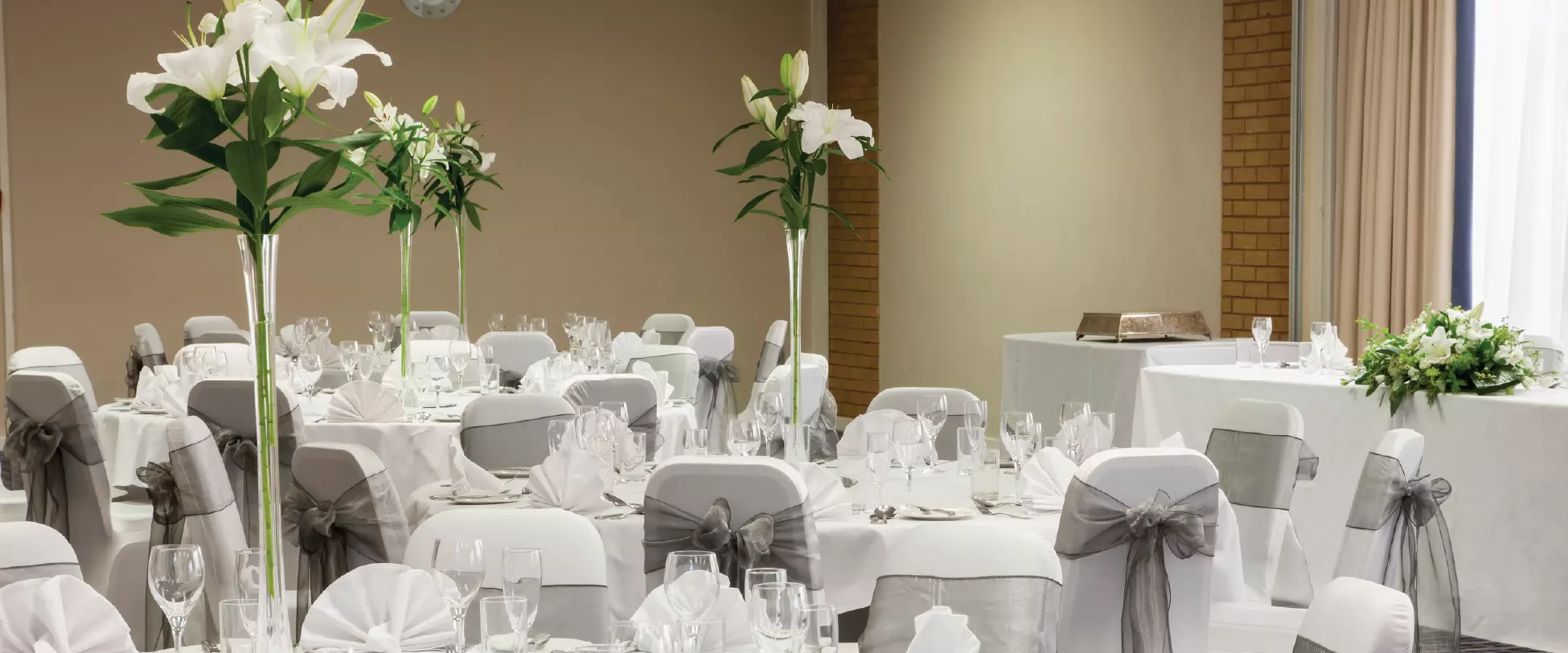 Tables set up for a wedding breakfast with white table and chair covers, silver ribbons and tall floral centrepieces..